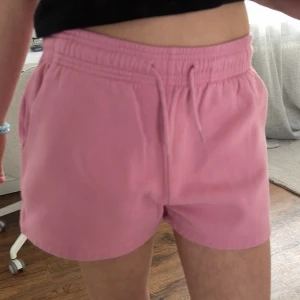 Rosa shorts med snörning från 157 - Säljer ett par ljusrosa shorts från 157 med elastisk midja och snörning framtill. Shortsen har en relaxed passform och är tillverkade i mjuk bomull, perfekta för varma dagar. Klassisk design utan mönster och med fickor på sidorna.