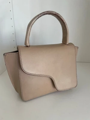 Beige handväska från ATP Atelier - Stilren beige handväska från ATP Atelier i slätt läder. Väskan har ett topphandtag och en unik asymmetrisk lockdesign. Tillverkad i Italien och perfekt för dig som gillar minimalistisk och modern stil.