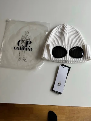 mössa C.P. Company - Säljer en vit ribbstickad mössa från C.P. Company med de ikoniska svarta goggles framtill. Mössan har en tight passform och är tillverkad i mjukt material som håller dig varm. Perfekt accessoar för dig som vill sticka ut och addera streetwear-vibe till din outfit.