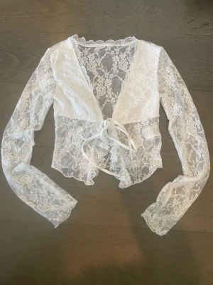 Vit spetsbolero med knyt - Säljer en superfin vit bolerotopp i spets med långa ärmar och v-ringning. Toppen har knyt framtill och är helt genombruten med blommigt spetsmönster. Perfekt för att lägga till en extra detalj till din outfit. Köpt sacond hand