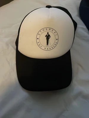 Svartvit keps från Ethereal - Snygg svartvit keps från Ethereal med tryckt logga framtill. Framsidan är vit med svart motiv och text, medan skärmen och baksidan är svarta. Baksidan har meshmaterial för extra ventilation och justerbar passform. Perfekt för en trendig och avslappnad stil.