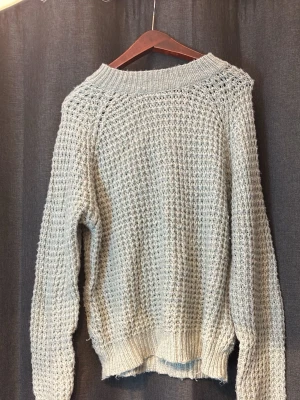 Ljusgrå stickad tröja med struktur - Ljusgrå stickad tröja med grov struktur och bred ribbad mudd vid ärmslut och nederkant. Tröjan har en avslappnad passform och rund halsringning. Perfekt för lager på lager och mysiga dagar.