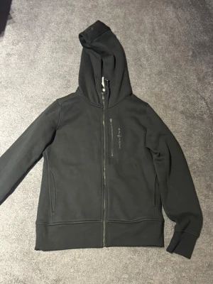 Svart zip hoodie från Sail Racing - Snygg svart hoodie från Sail Racing med dragkedja framtill och broderad logga på bröstet. Modellen har huva med dragsko och två fickor. Tillverkad i mjukt och bekvämt material som passar perfekt till chill eller streetwear.