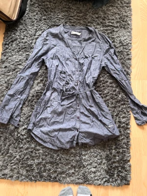 Grå klänning med knytband - Säljer en grå långärmad skjorta från Hunkydory med knappar framtill och ett matchande knytband i midjan. Skjortan har en avslappnad passform och mjukt material som känns skönt mot huden. Perfekt för en cool och enkel look.