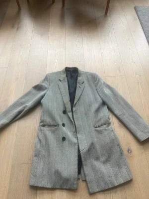 Tiger of Sweden rock med grårandigt mönster - Snygg grå rock med diskreta vertikala ränder och tre knappar framtill. Rocken har två fickor med lock och en klassisk krage. Fodrad insida och längre modell som ger en stilren siluett. Perfekt för dig som vill ha en tidlös och trendig look. Kan diskuteras pris