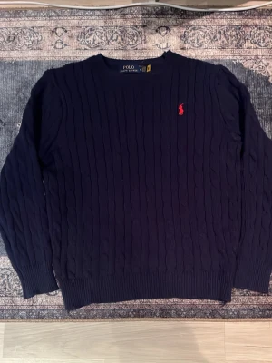 Mörkblå kabelstickad tröja Polo Ralph Lauren - Kabelstickad mörkblå tröja från Polo Ralph Lauren med rund halsringning och röd broderad logga på bröstet. Tröjan har långa ärmar och ribbade muddar vid ärmslut och nederkant. Perfekt för dig som gillar klassisk och stilren look.
