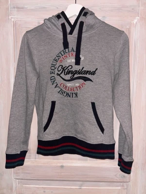 Grå hoodie från Kingsland - Snygg grå hoodie från Kingsland med broderad text och logga på bröstet. Muddar och nederkant har svarta, röda och gröna ränder. Huva med svarta snören och känguruficka framtill. Perfekt för en avslappnad och sportig stil. (Nypris 1099kr, bara att höra av sig vid intresse, pris kan regleras vid smidig affär) Hoodien är i nyskick o knappt använd💞