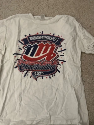 Vit t-shirt Norgesmesterskapet 2025 - Vit t-shirt med stort tryck på framsidan i rött, blått och vitt. Texten på tröjan lyder 'NORGESMESTERSKAPET', 'Performance cheer & Cheerleading 2025'. Perfekt för dig som gillar cheerleading och sportig stil.
