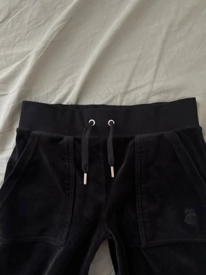 Juciy couture byxor - Säljer ett par svarta mjukisbyxor i mjuk velour med bred resår i midjan och snörning framtill. Byxorna har stora fickor fram och bakfickor med knappdetaljer. Perfekta för en chill och avslappnad stil.