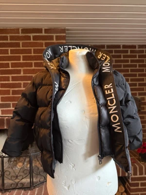 Svart pufferjacka från Moncler - Säljer en svart pufferjacka från Moncler med hög krage och tydlig logotyp längs dragkedjan. Jackan har en klassisk puffad design och är tillverkad i glansigt material som ger en exklusiv känsla. Perfekt för kalla vinterdagar och riktigt snygg till streetwear-stilen.