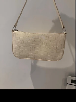 Beige axelremsväska i crocopräglat skinn - Snygg beige handväska med axelrem och crocopräglat mönster. Väskan har en avlång, rektangulär form och stängs med dragkedja upptill. Perfekt för dig som gillar stilrena accessoarer med trendig touch.