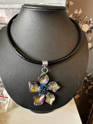 Färgstarkt blomhalsband i metall - Unikt halsband med ett stort, färgglatt blomformat hänge i metall. Hänget har regnbågsskimrande nyanser i blått, gult, rosa och lila samt en detaljerad mitt. Svart läderrem med tre silverfärgade ringar vid hänget. Perfekt statement piece för dig som vill sticka ut.