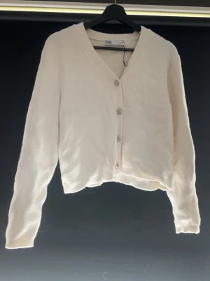 Vit kofta från Zara med pärlknappar - Snygg vit kofta från Zara med v-ringning och tre dekorativa pärlknappar framtill. Koftan har lång ärm, en bröstficka och är gjord i ett mjukt, fluffigt material. Perfekt att slänga över en topp för en chill look.