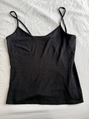 Svart linnetopp med smala axelband - Enkel och stilren svart linnetopp med smala axelband. Toppen har en rak passform och ärmlös design, perfekt att matcha med jeans eller kjol. Materialet känns mjukt och stretchigt, vilket gör den bekväm att bära hela dagen.