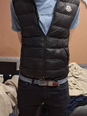 Svart dunväst från Moncler - Snygg svart dunväst från Moncler med klassisk quiltad design och dragkedja framtill. Västen har två sidofickor med dragkedja och Monclers ikoniska logga på bröstet. Perfekt att slänga över en hoodie eller t-shirt för en trendig look.