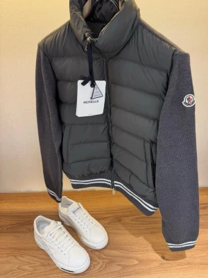 Moncler grå dunväst med stickade ärmar - Snygg grå dunväst från Moncler med stickade mörkgrå ärmar och vita ränder vid muddar och nederkant. Västen har dragkedja framtill, hög krage och Moncler-logga på ärmen. Perfekt för dig som vill ha en sportig och trendig look. Material: dun och stickad textil.