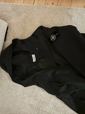 Svart vindjacka från Stone Island - Säljer en svart vindjacka från Stone Island med ikonisk patch på vänster ärm. Jackan har dragkedja framtill, huva och en stilren, sportig look. Perfekt för dig som gillar streetwear och vill ha något lätt och snyggt till vardagen. Jag som på bilden är 170cm lång och sitter bra men lite stor runt armarna