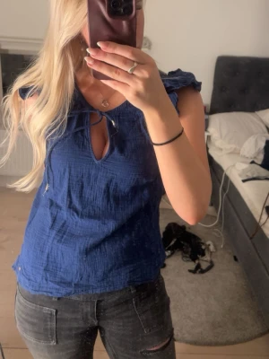 Blå blus med knyt och volangärmar - Snygg blå blus med knyt i halsen och liten öppning framtill. Toppen har volangdetaljer vid axlarna och är ärmlös. Tillverkad i ett lätt, strukturerat tyg som känns luftigt och skönt. Perfekt för dig som vill ha en trendig och bekväm topp.