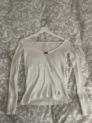 Vit ribbad topp från Odd Molly - Supersöt vit långärmad topp från Odd Molly i ribbat material. Djup v-ringning med en liten vinröd rosett framtill och diskret textdetalj på insidan. Figurnära passform och mjukt tyg som känns skönt mot huden. Storlek 0 alltså XS