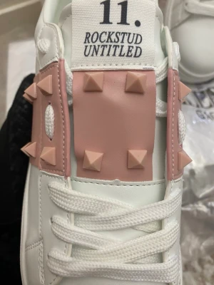 Valentino Rockstud Untitled sneakers - Snygga vita sneakers från Valentino med ljusrosa detaljer och ikoniska pyramidformade nitar. Modellen har en chunky vit sula och breda vita skosnören. Ovandelen är i läder och skon har en modern, trendig look med coola detaljer på sidan och tungan.