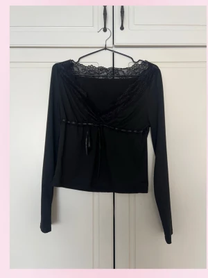 Svart omlottopp med spetsdetaljer - 💓Snygg svart omlottopp med långa ärmar och spetsdetaljer längs ringningen. Toppen har en dekorativ knytning under bysten och är tillverkad i ett mjukt, stretchigt material. Perfekt för dig som gillar en feminin och trendig stil.💕