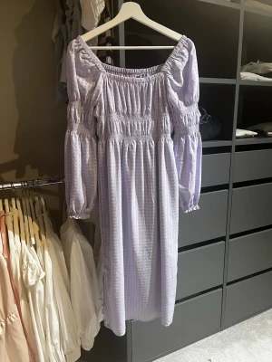 Ljuslila offshoulder klänning - Söt ljuslila klänning med offshoulder och långa puffärmar. Klänningen har smockdetaljer upptill och vid ärmarna samt ett diskret rutigt mönster. Perfekt för sommarens alla tillfällen!
