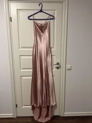 Lång rosa satinklänning med smala band - Elegant långklänning i skimrande rosa satin med tunna axelband och snygg omlott-detalj i ryggen. Klänningen har en mjukt draperad urringning och faller fint längs kroppen. Perfekt för speciella tillfällen när du vill känna dig extra glam.