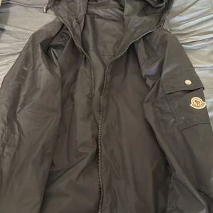 Svart vindjacka från Moncler - Snygg svart vindjacka från Moncler med huva och dragkedja framtill. Jackan har en ficka med knapp på ärmen och Moncler-logga i tyg på armen. Tillverkad i lätt, glansigt material som står emot vind. Perfekt för dig som vill ha en stilren och funktionell jacka.