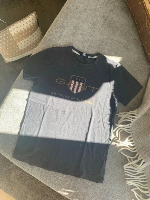 Mörkgrå GANT t-shirt med tryck - Snygg mörkgrå t-shirt från GANT med klassiskt tryck framtill och rund hals. T-shirten har korta ärmar och är tillverkad i mjuk bomull, perfekt för en avslappnad och sportig stil. Storlek 146-152 cm, 11-14 år.