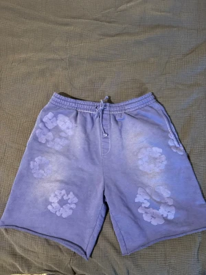 Denim Tears shorts - Säljer ett par lila sweatshorts med dragsko i midjan och vita blommönster tryckta över hela plagget. Shortsen har avslappnad passform och är gjorda i mjukt sweatshirtmaterial, perfekta för chill dagar.