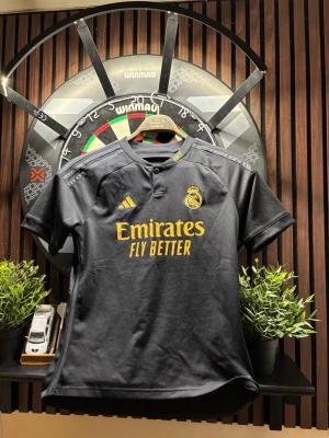 Real Madrid 2023/2024 tredjeställ  - Snygg svart Real Madrid matchtröja från Adidas med guldfärgade detaljer och klubbmärke på bröstet. Tröjan har korta ärmar, rund hals med knapp och 'Emirates Fly Better' tryckt på framsidan. Tillverkad i lätt och ventilerande material. Köpte denna ny på unisport för 2 år sedan och är knappt använd, äkta såklart! Storlek 164 men sitter som Xs/S