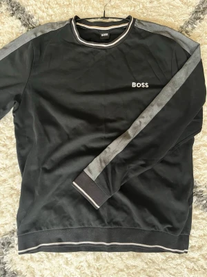 Svart sweatshirt från BOSS - Svart sweatshirt från BOSS med gråa detaljer längs ärmarna och vita ränder vid krage, ärmslut och nederkant. Tröjan har rund halsringning och BOSS-logga broderad på bröstet. Perfekt för en avslappnad och stilren look. Storlek L men massor bättre som Stor S eller M…