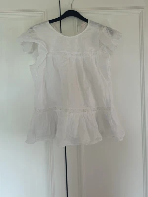 Vit blus med volang från Gina Tricot - Söt vit blus från Gina Tricot med volangkant nedtill och korta puffärmar. Blusen har rund halsringning och tunnt, luftigt tyg som passar perfekt till sommaren. Enkel och stilren design som är lätt att matcha.