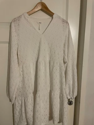 Vit spetsklänning  - Söt vit klänning med v-ringning och långa ärmar. Klänningen har volangdetaljer och är tillverkad i spets med blommigt mönster. Perfekt för studenten. 