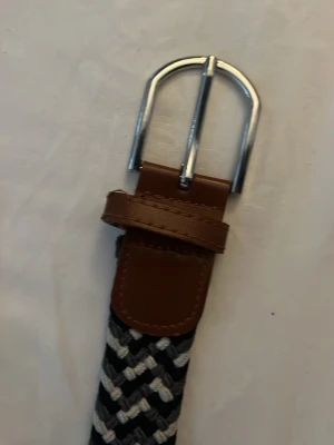 Randigt flätat bälte med läderdetalj - Snyggt flätat bälte i svart och vitt med randigt mönster. Spännet är i silverfärgad metall och bältet har bruna läderdetaljer vid spännet. Perfekt accessoar för att lyfta din outfit.