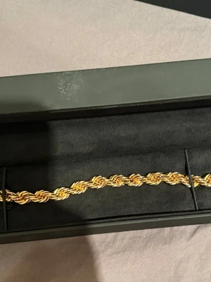 Flätat guldarmband från Kejsar - Snyggt flätat armband från Kejsar i äkta 18k guld platerad. Armbandet har en tvinnad design som ger en exklusiv känsla och levereras i en elegant svart ask med Kejsar-logga i gult. Perfekt accessoar för att lyfta din stil.