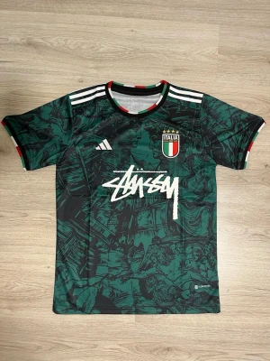 ItalienXstussy fotbollströja perfekt till sommaren - Grön och svart fotbollströja från Adidas i samarbete med Stüssy och Italien. Tröjan har vita Adidas-ränder på axlarna, Italiens landslagsemblem på bröstet och Stüssy-logga framtill. Kragen har italienska flaggans färger. Tillverkad i lätt funktionsmaterial.