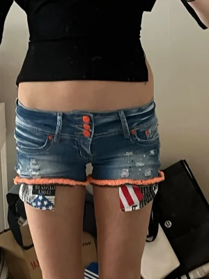 Blå jeansshorts med orange detaljer - Unika blå jeansshorts med orange knappar och fransig kant. Shortsens uppvik har färgglada tygdetaljer med mönster och slitningar framtill för en edgy look. Passformen är tight och modellen är lågmidjad. Perfekta för sommaren och festivaler.