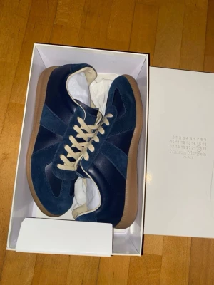 Blå Maison Margiela sneakers - Snygga blå sneakers från Maison Margiela med ovandel i mocka och läder, beigea skosnören och klassisk gummisula. Skorna har en stilren och tidlös design som passar perfekt till streetwear och casual outfits.
