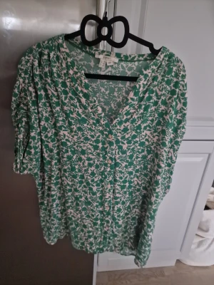 Grönblommig blus från Esprit  - Snygg blus från Esprit i grönt och vitt med blommigt mönster. Blusen har v-ringning och trekvartsärm med lätt puff och rynkdetaljer. Tillverkad i ett mjukt, luftigt tyg som känns skönt mot huden. Perfekt för dig som gillar en avslappnad men trendig stil.