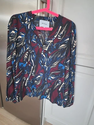 Mönstrad blus från Minus - Snygg blus från Minus med abstrakt mönster i vinrött, blått, svart.. Blusen har V-ringning och långa ärmar med lätt puff vid axlarna. 