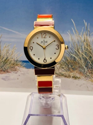 Guldig klocka från In Zone - Snygg klocka från In Zone med guldfärgat armband och boett. Armbandet har inslag av rött, orange och beige i blockfärg. Urtavlan är vit med guldiga detaljer och siffror. Perfekt accessoar för dig som vill sticka ut lite extra.