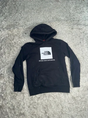 Svart hoodie från The North Face - Säljer en svart hoodie från The North Face med stor logga och texten 'Never Stop Exploring' på bröstet. Klassisk huva, ribbade muddar och fickor framtill. Perfekt för dig som gillar streetwear och vill ha en stilren look. Varan är i bra skick, för fler bilder/frågor skriv privat