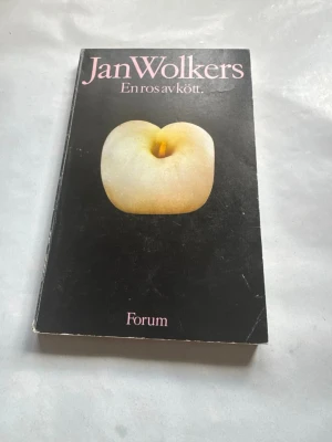 En ros av kött av Jan Wolkers - Romanen 'En ros av kött' av Jan Wolkers har ett svart mjukt omslag med en konstnärlig bild av en ljusgul persika och rosa text. Boken är i pocketformat och utgiven av Forum. Perfekt för dig som gillar modern skönlitteratur och unika omslag.