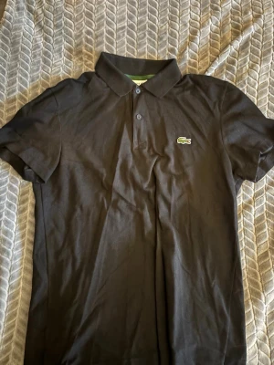 Svart pikétröja från Lacoste - Klassisk svart pikétröja från Lacoste med den ikoniska gröna krokodilloggan på bröstet. Tröjan har korta ärmar, krage och knäppning med två knappar framtill. Perfekt för en clean och stilren look.
