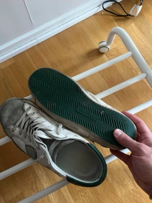 Philippe Model sneakers - Säljer ett par vita sneakers med grön yttersula och detaljer i svart och grönt på hälen. Skorna har klassisk låg profil, vita skosnören och är tillverkade i läder. Perfekta för dig som gillar en clean och enkel stil.