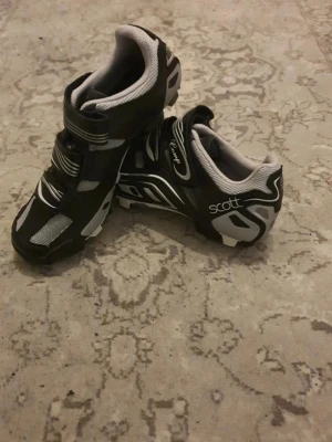 Svarta cykelskor från Scott - Säljer ett par svarta cykelskor från Scott med vita och grå detaljer. Skorna har robust sula med SPD-fäste, kardborreband och meshpaneler för ventilation. Perfekta för MTB eller spinning och ger bra grepp och stöd under cyklingen.