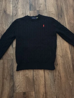 Svart kabelstickad tröja Polo Ralph Lauren - Kabelstickad svart tröja från Polo Ralph Lauren med rund halsringning och röd broderad logga på bröstet. Klassisk passform och ribbade muddar vid ärmslut och nederkant. Perfekt för dig som gillar stilrena och tidlösa plagg.