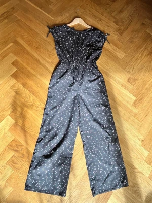 Svart blommig jumpsuit med vida ben - Snygg svart jumpsuit med små vita och rosa blommor. Modellen har vida ben, resår i midjan och korta ärmar med knytband på axlarna. Perfekt för dig som gillar en avslappnad men trendig stil. Materialet känns mjukt och lätt. Perfekt i sommar