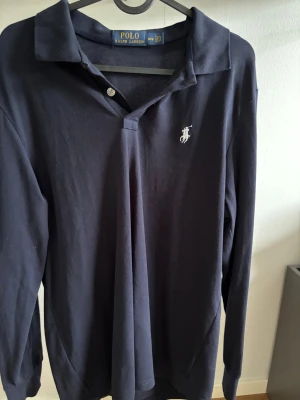 Mörkblå långärmad skjorta Polo Ralph Lauren - Klassisk mörkblå långärmad skjorta från Polo Ralph Lauren med vit broderad logga på bröstet. Skjortan har krage, knappar vid halsen och är tillverkad i mjuk bomull. Perfekt för en stilren och avslappnad look.